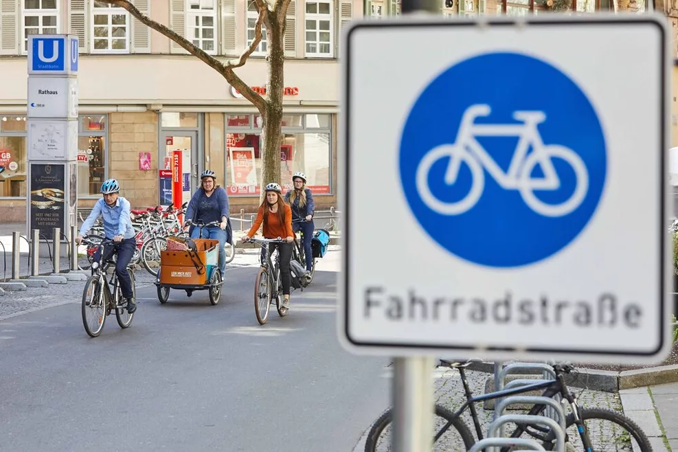 Verkehrsschild Fahrradstraße. Hinter dem Schild sind ein paar Fahrradfahrende zu sehen.